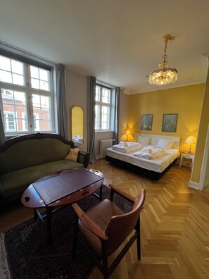 Une chambre d’hôtel avec un lit, un canapé, une table et une chaise. Il y a deux fenêtres avec des rideaux, un radiateur et un lustre.