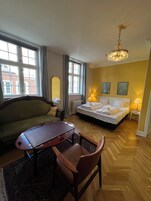 Habitación de hotel con cama, sofá, mesa y una silla. Hay dos ventanas con cortinas, un radiador y un candelabro.