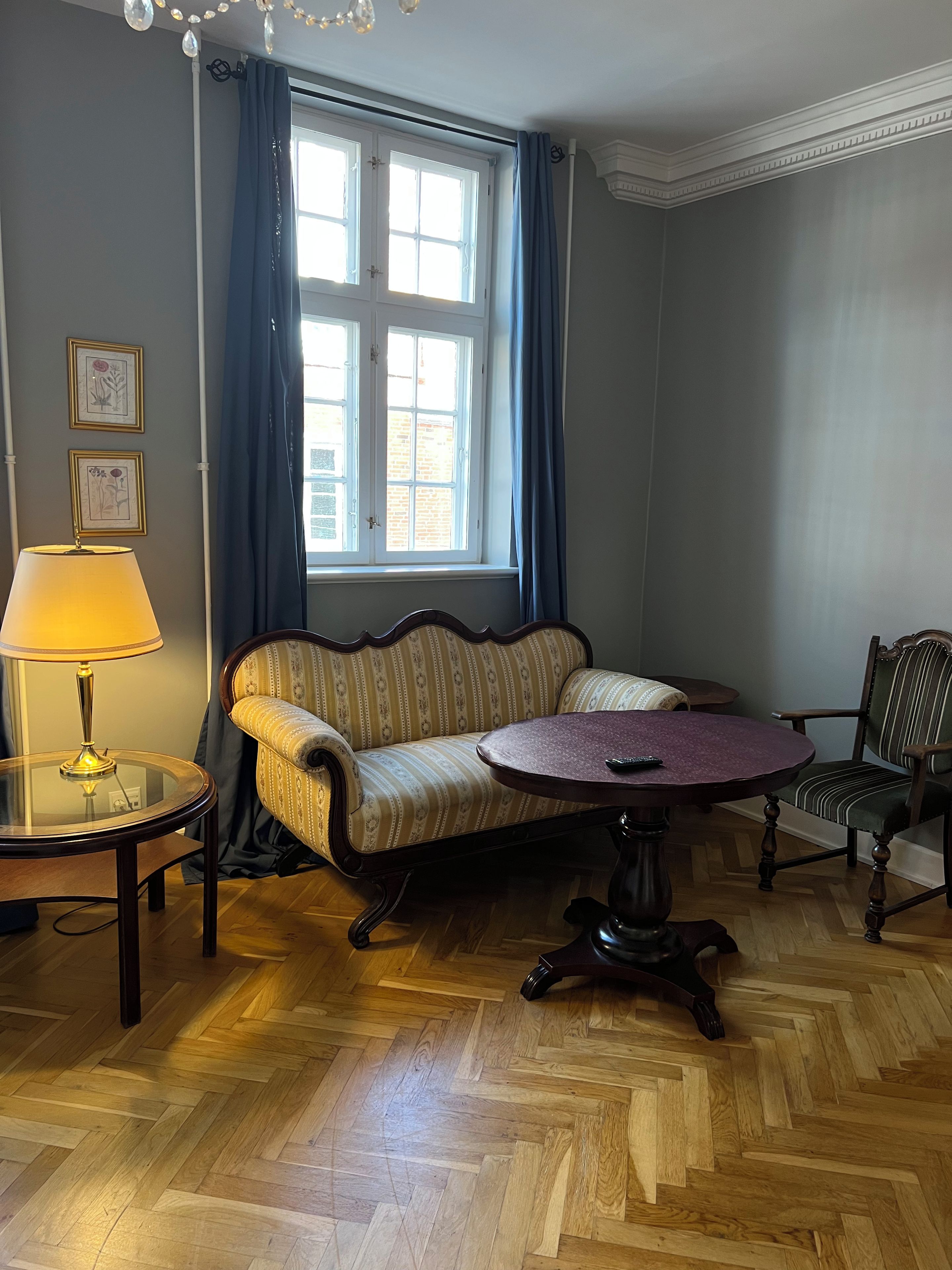 Una habitación con un sofá, una mesa redonda, una silla, una lámpara y un ventanal con cortinas.