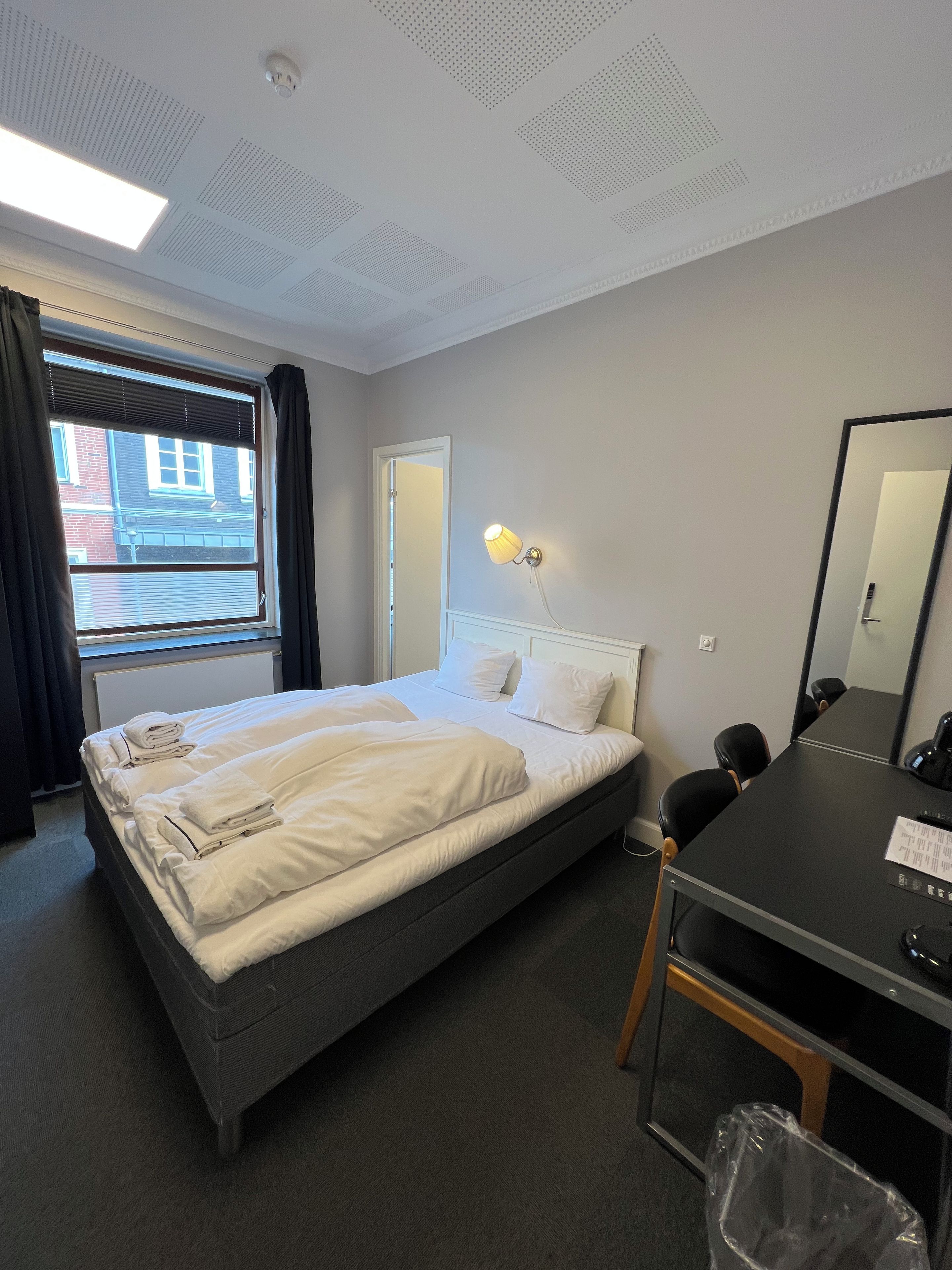 Habitación de hotel con cama, escritorio, silla, espejo y ventana con cortinas.