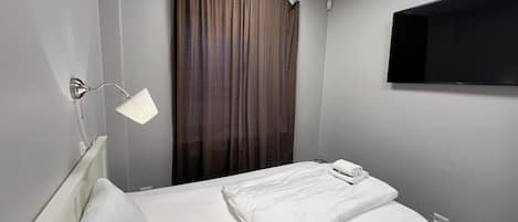 A hotel room with a single bed, a bedside lamp, a flat-screen TV mounted on the wall, and a window with curtains.