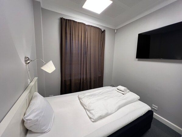 Habitación de hotel con cama individual, lámpara de noche, televisor de pantalla plana en la pared y ventana con cortinas.