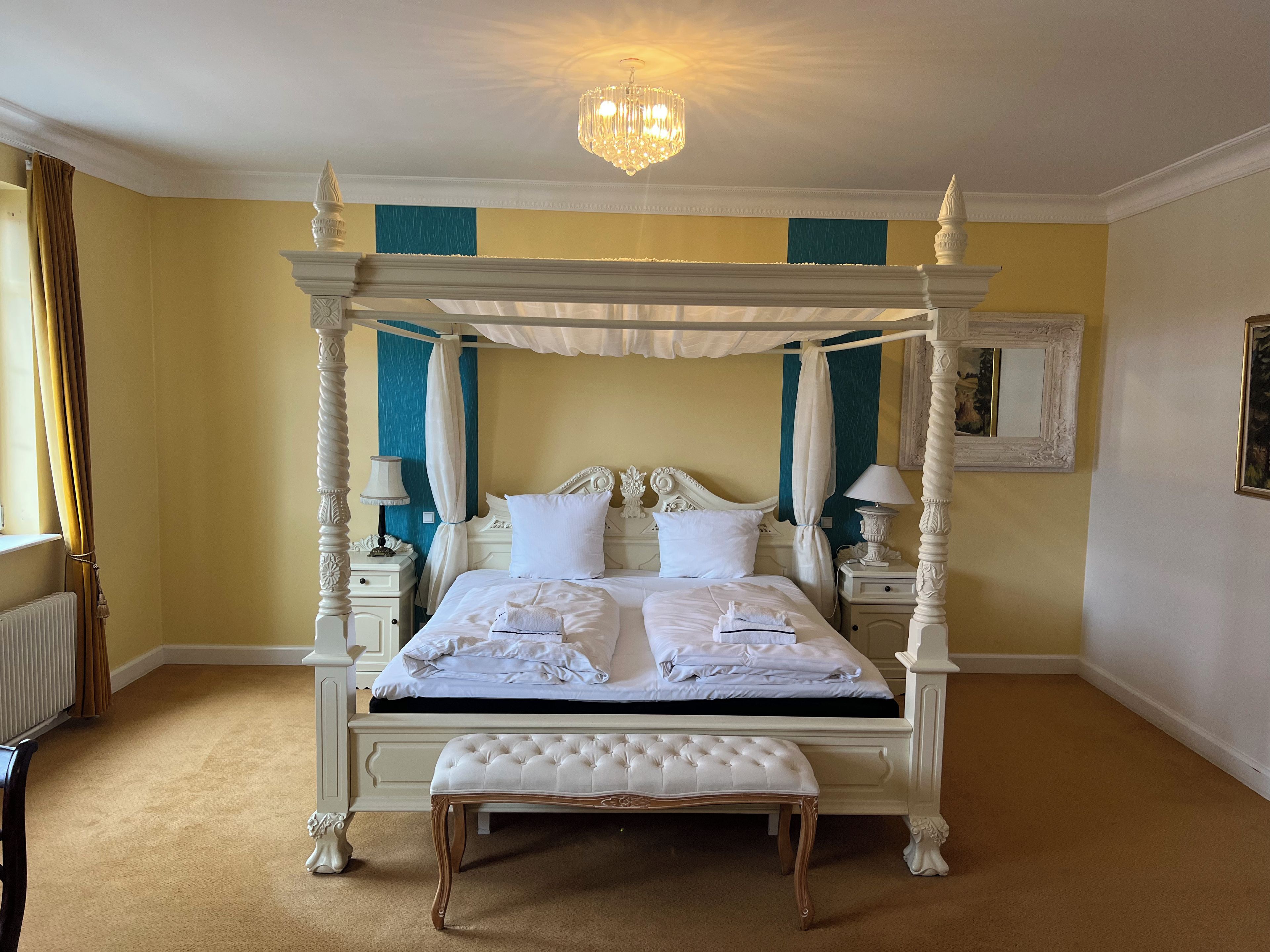 Ein Bett mit Baldachin, ausgestattet mit weißer Bettwäsche und einer passenden Bank, steht in einem Zimmer mit gelben Wänden und einem Heizkörper.