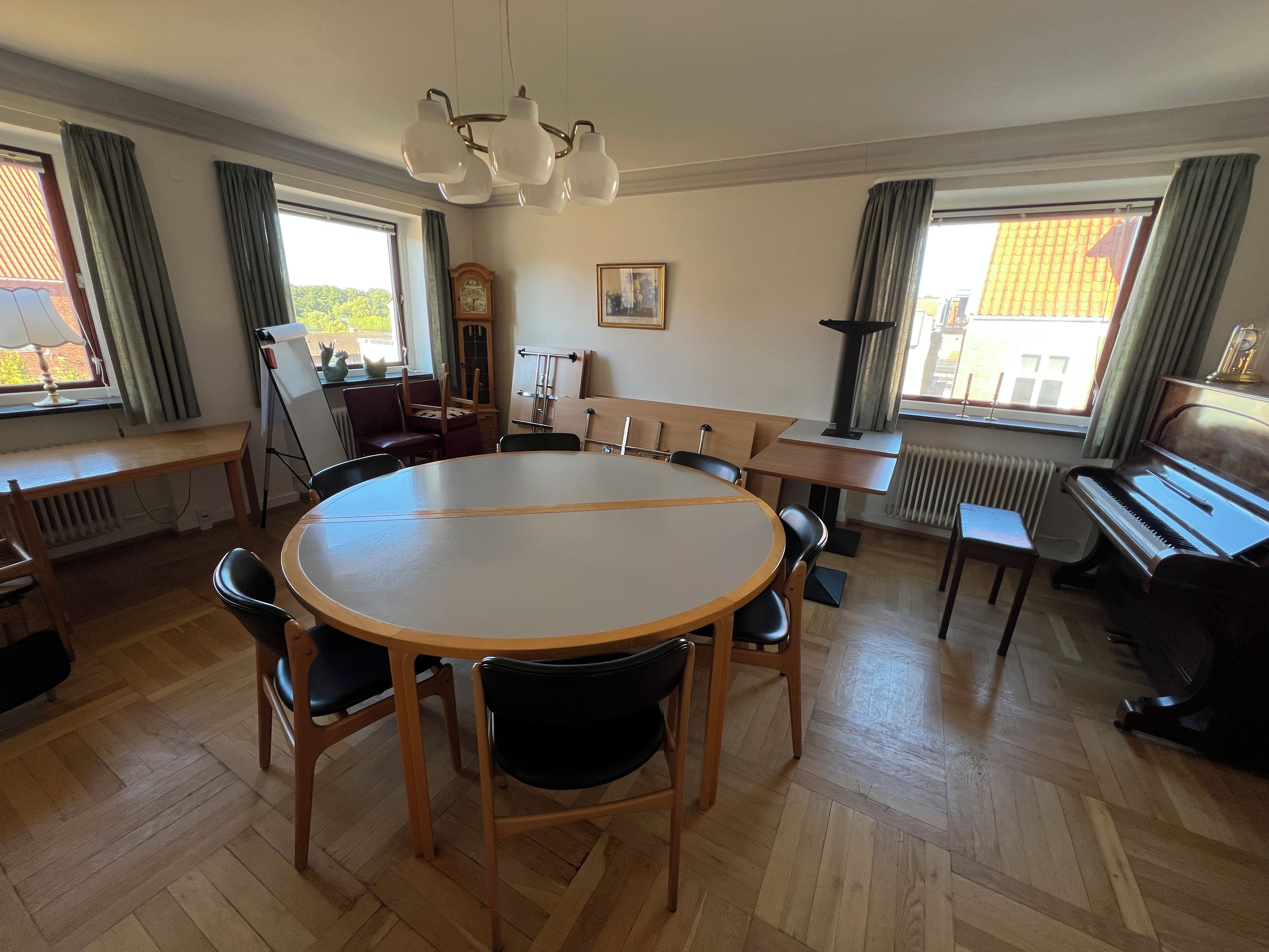 Una habitación amplia con una mesa redonda de madera rodeada de sillas, un piano y un radiador.