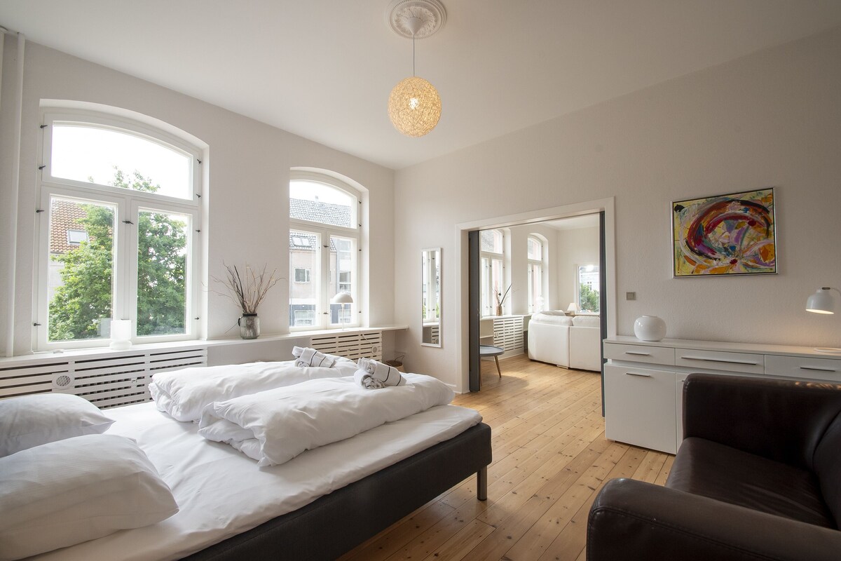 Una camera da letto moderna con un letto grande, una scrivania e una sedia. Diverse finestre offrono luce naturale e vista sull'esterno.
