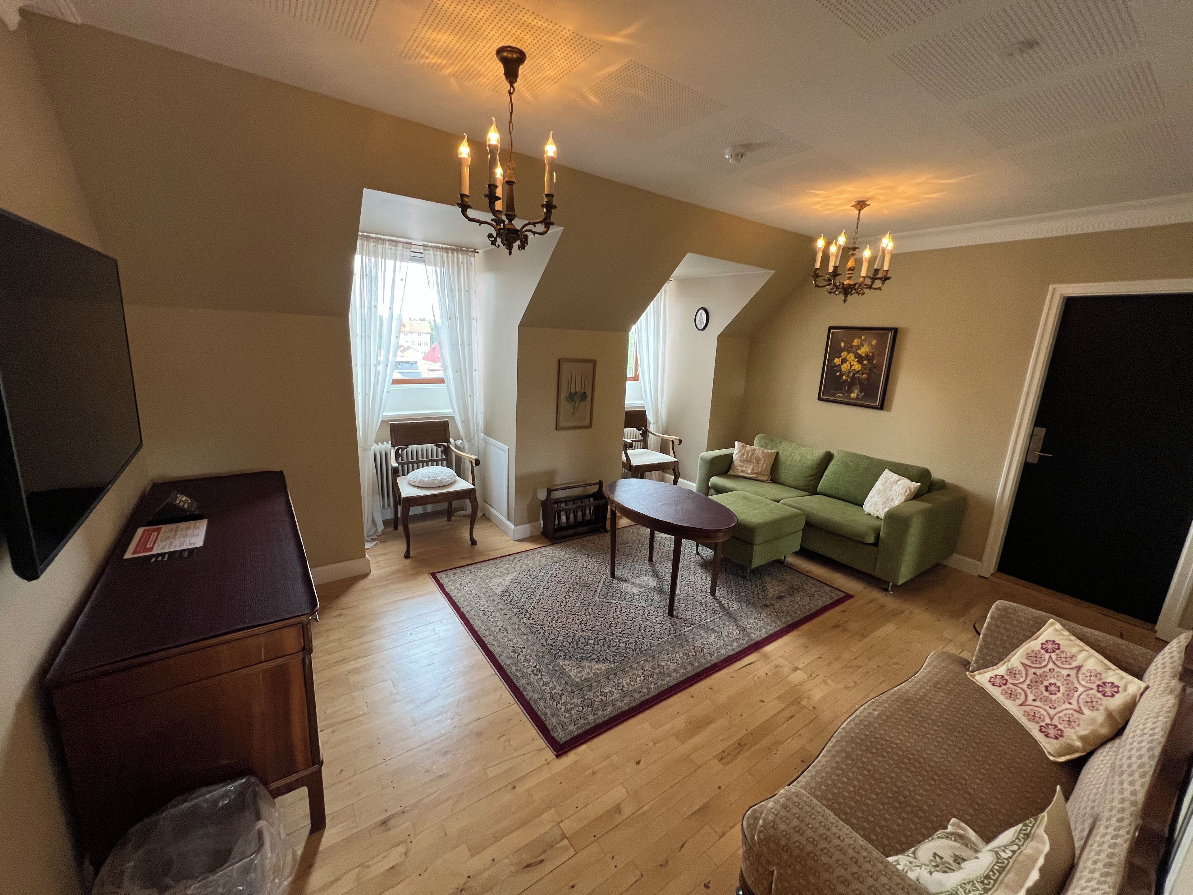 Ein Wohnzimmer mit einem grünen Sofa, einem braunen Sessel, einem Holztisch, einer Chandelüster und einem großen Fenster mit Vorhängen.