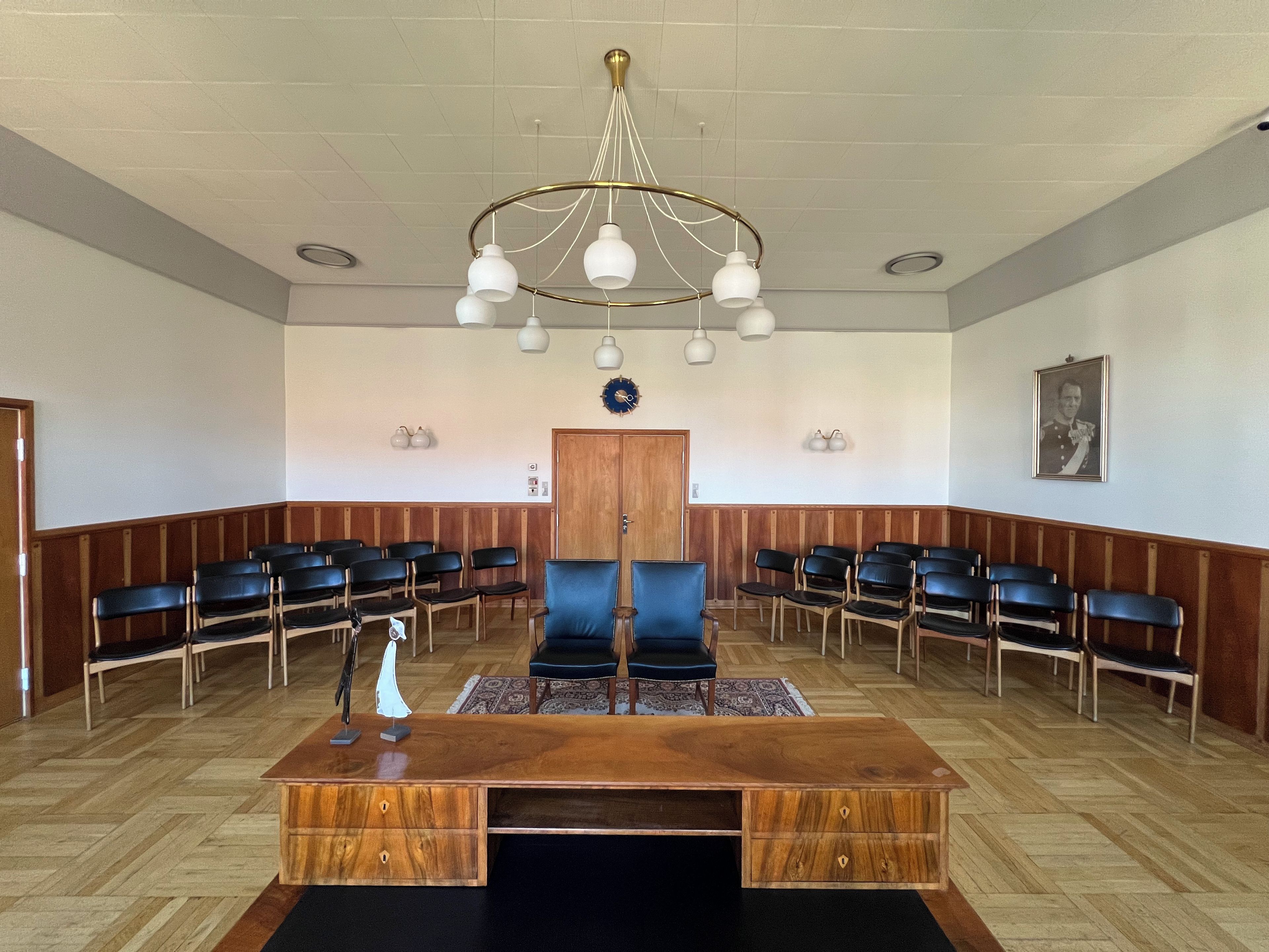 Un salón de conferencias con un escritorio de madera, sillas negras, una araña y un reloj en la pared.