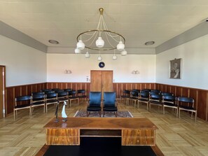 Une salle de conférence avec un bureau en bois, des chaises noires, un lustre et une horloge au mur.