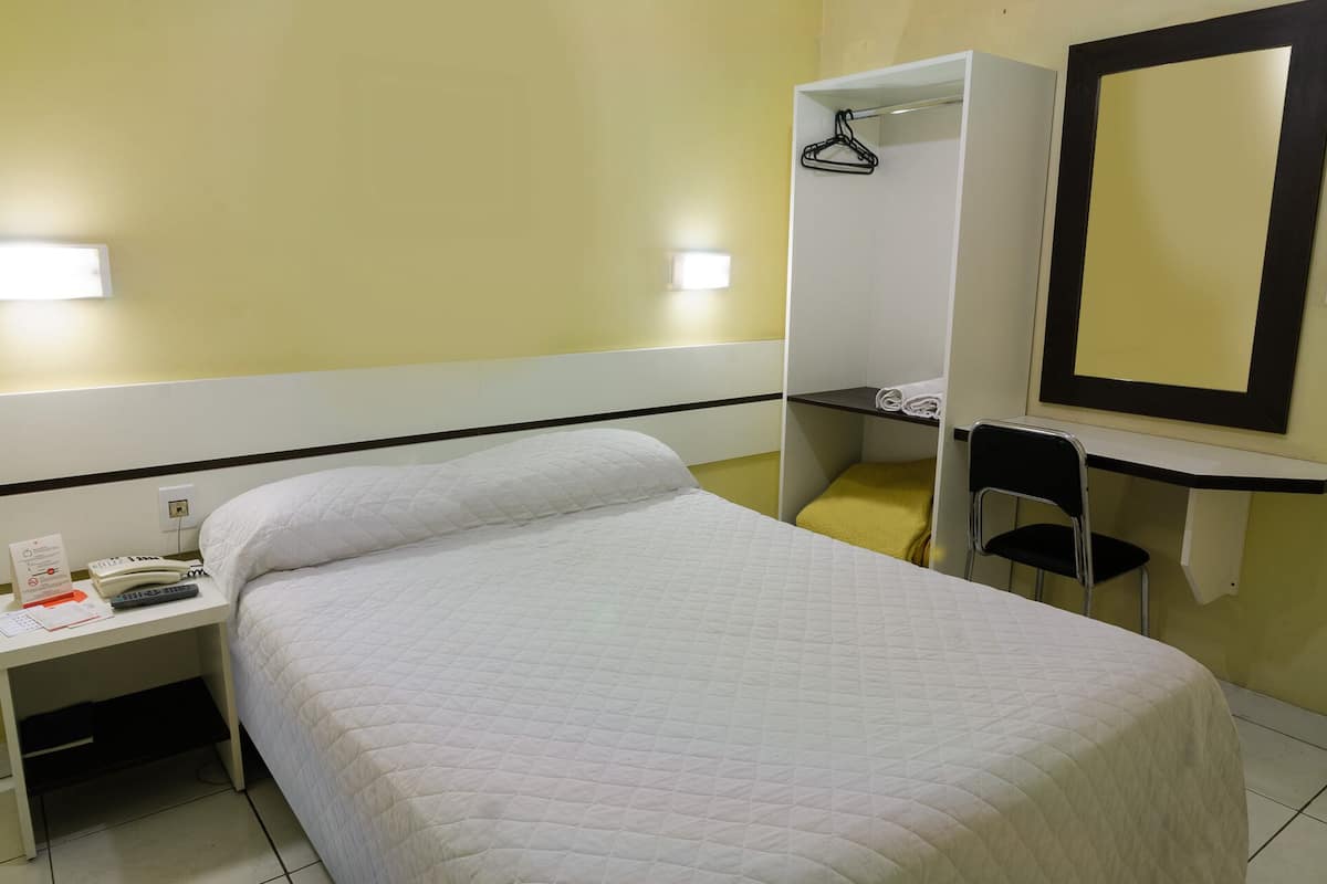 Ein Einzelbett mit weißer, gesteppter Decke, ein Schreibtisch mit Telefon und Unterlagen, ein Stuhl und ein Spiegel.