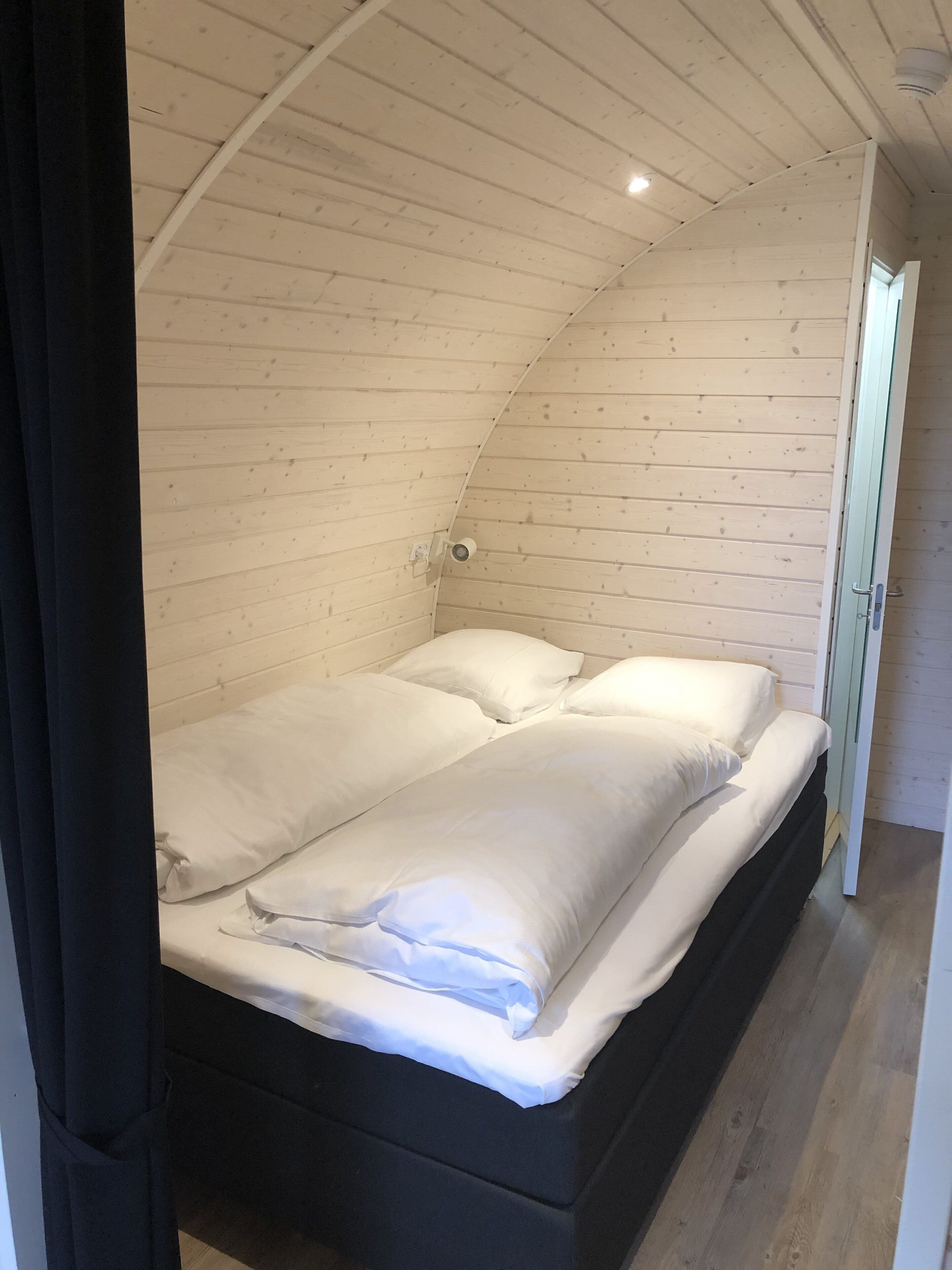Un dormitori petit i acollidor amb sostre de fusta inclinat, un llit individual amb roba de llit blanca i una porta visible que dona a una altra habitació.