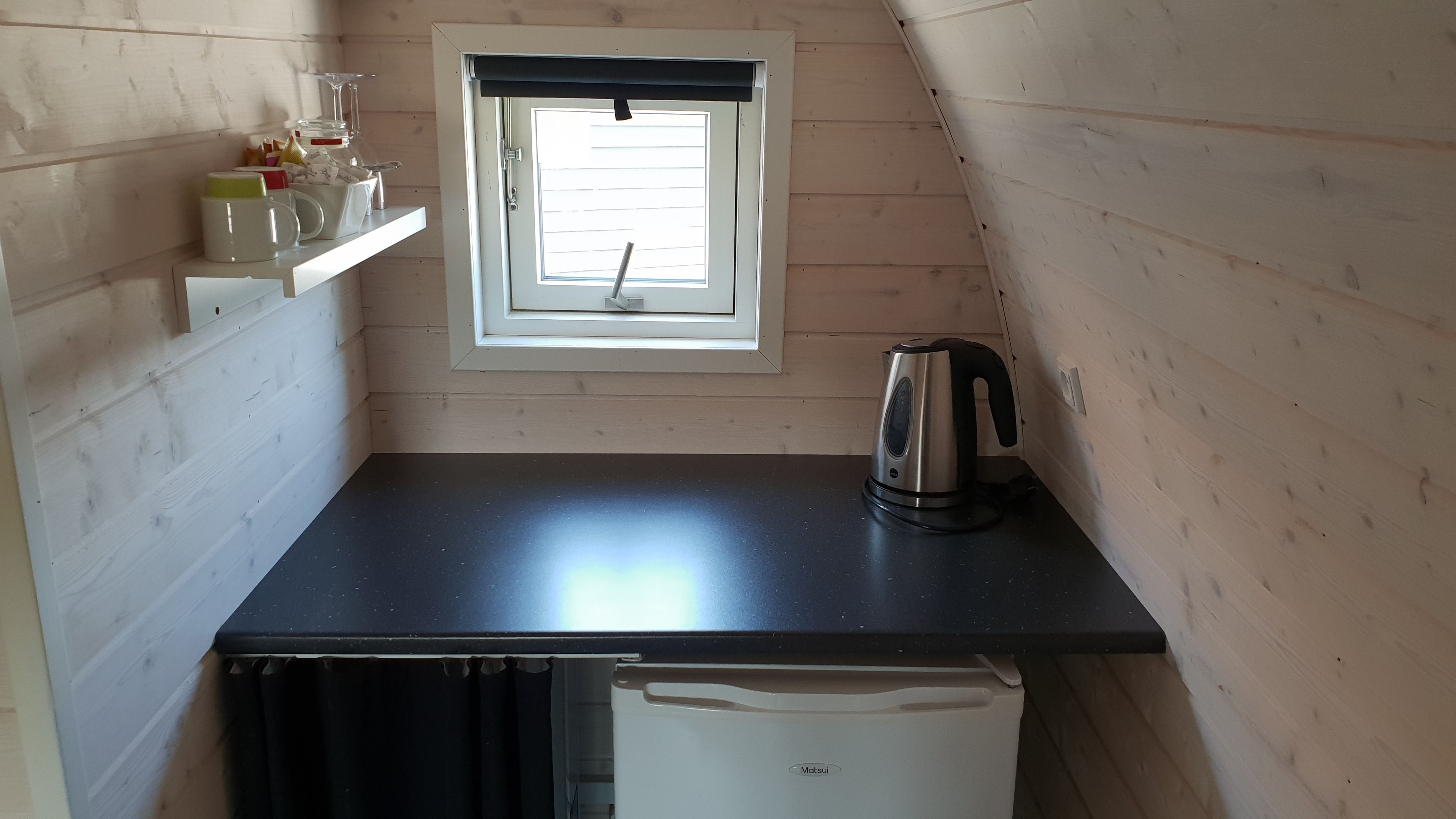 Una cocina compacta con encimera negra, hervidor, refrigerador y ventana con persianas.