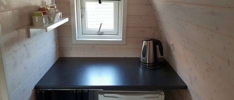 A compact kitchen with a black countertop, a kettle, a refrigerator, and a window with blinds.