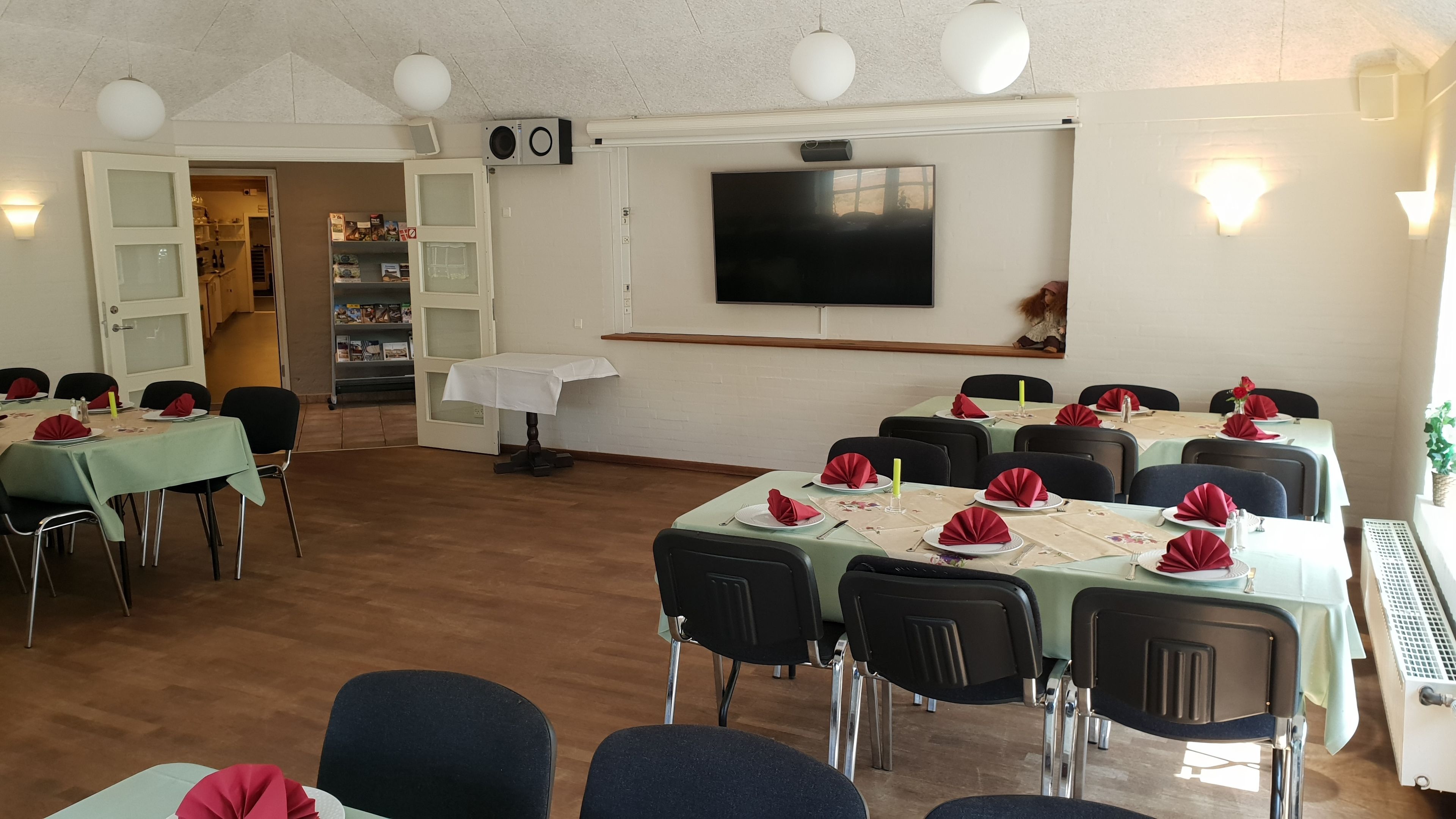 Un comedor con mesas dispuestas para varias personas, un televisor montado en la pared y una puerta que da a otra habitación.