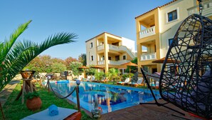 A pool area with lounge chairs, a hammock, and a building with balconies. - Garden Blue By Greoudis (Platanias)