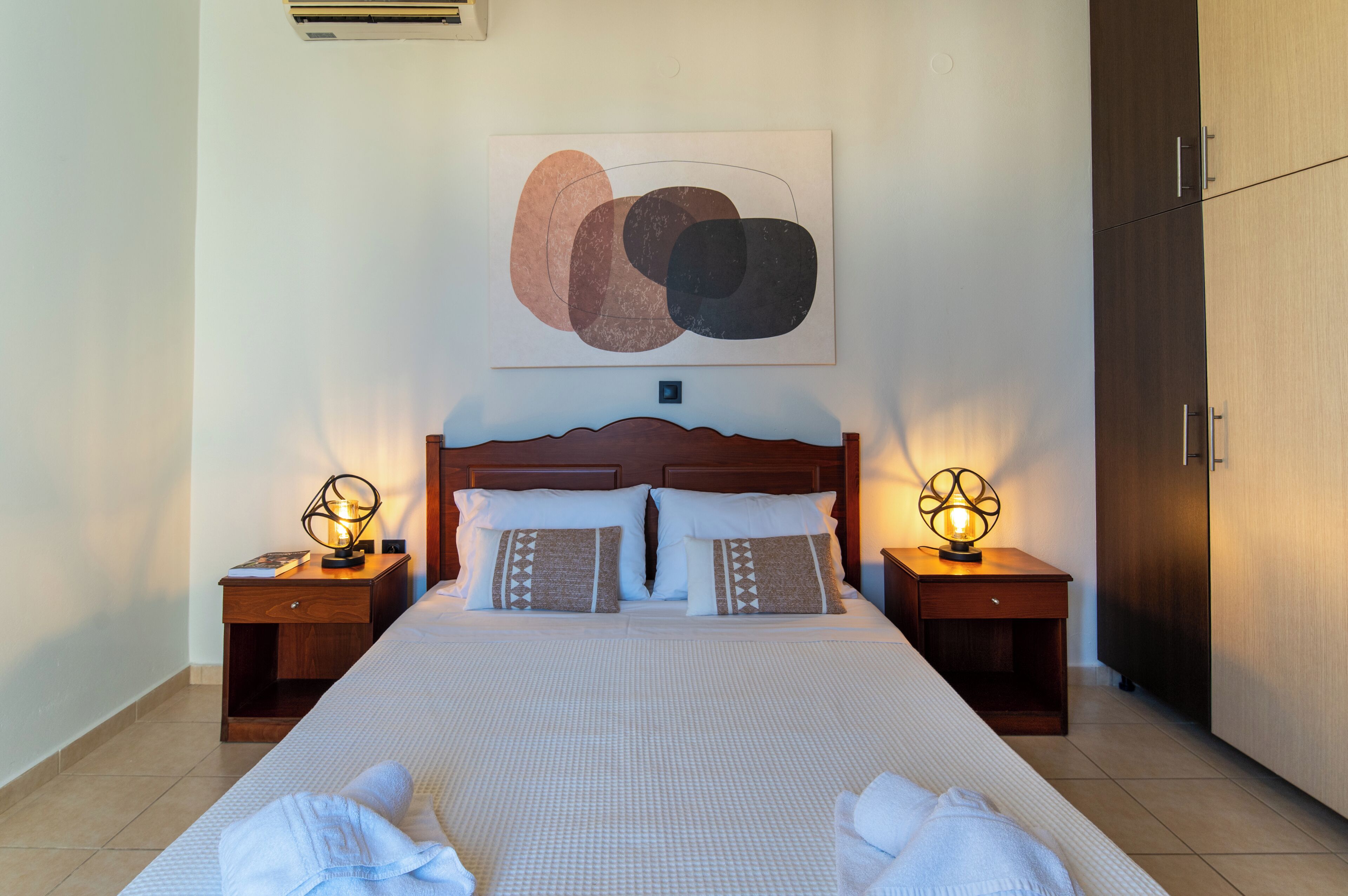 Un lit bien fait, avec deux oreillers, une lampe de chevet et un tableau abstrait accroché au mur.