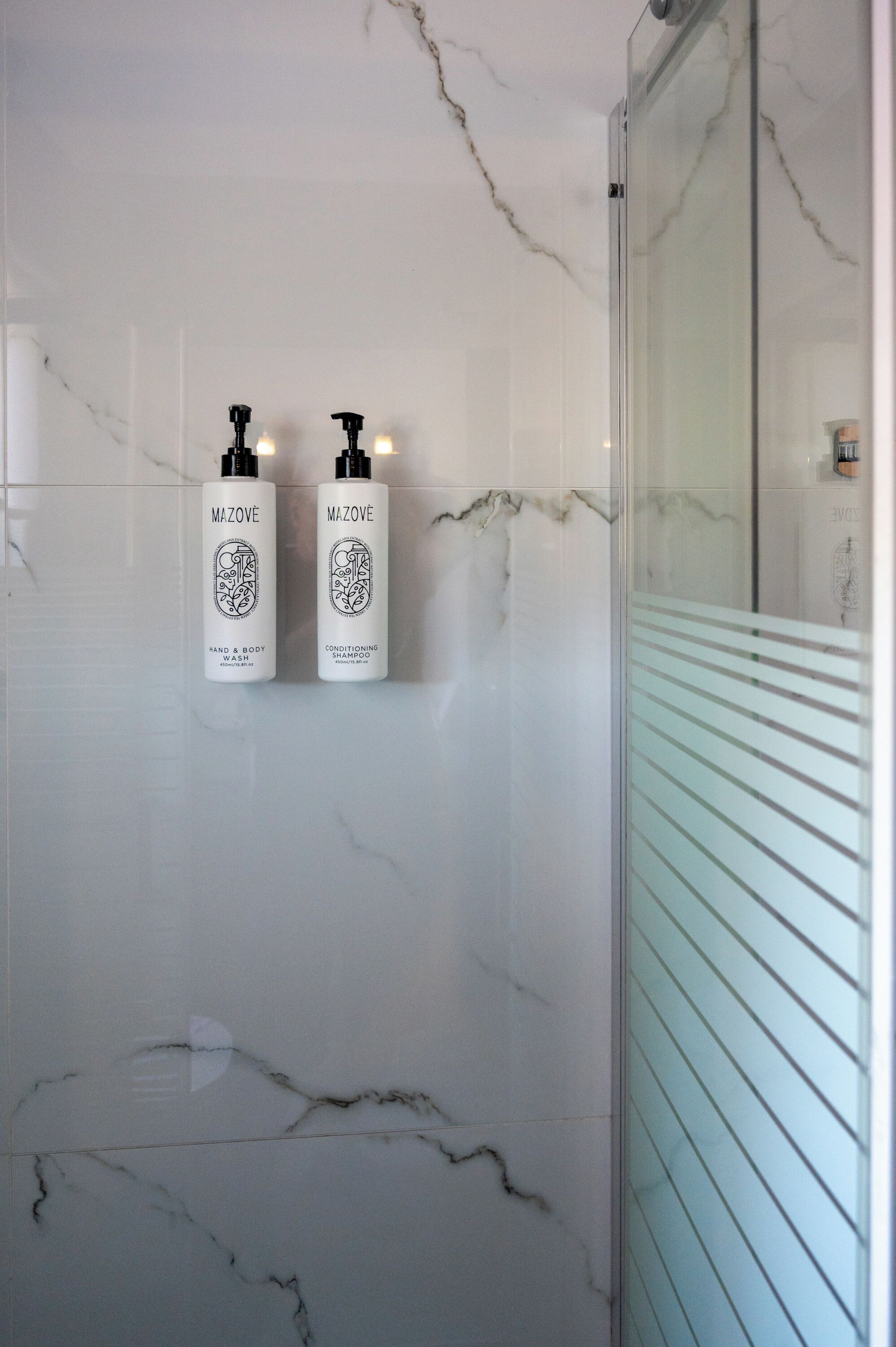 A bathroom with marble walls, two hand and body care products, and a glass shower enclosure.
