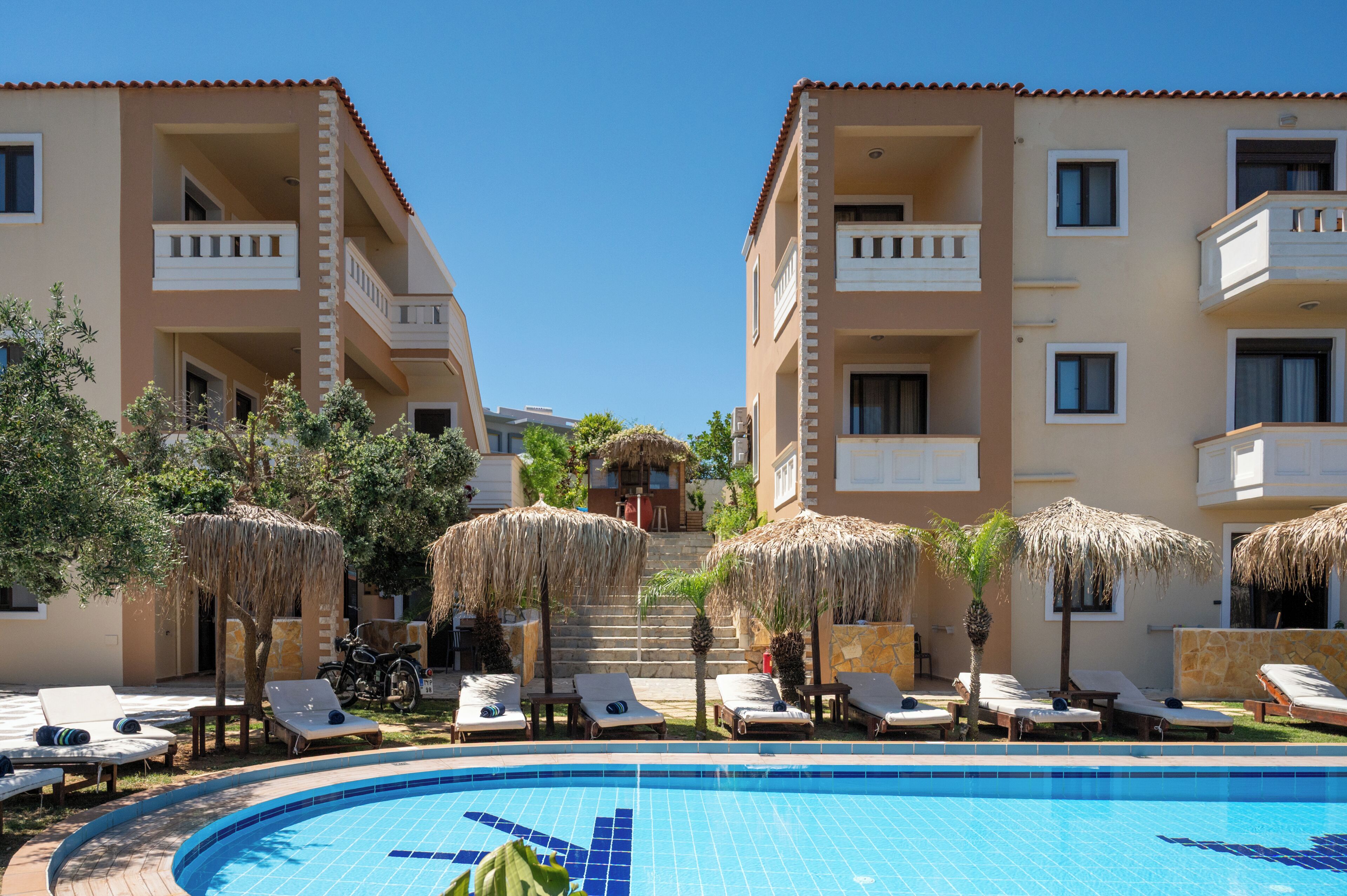 Un hotel con piscina, sillas de descanso y sombrillas de paja.