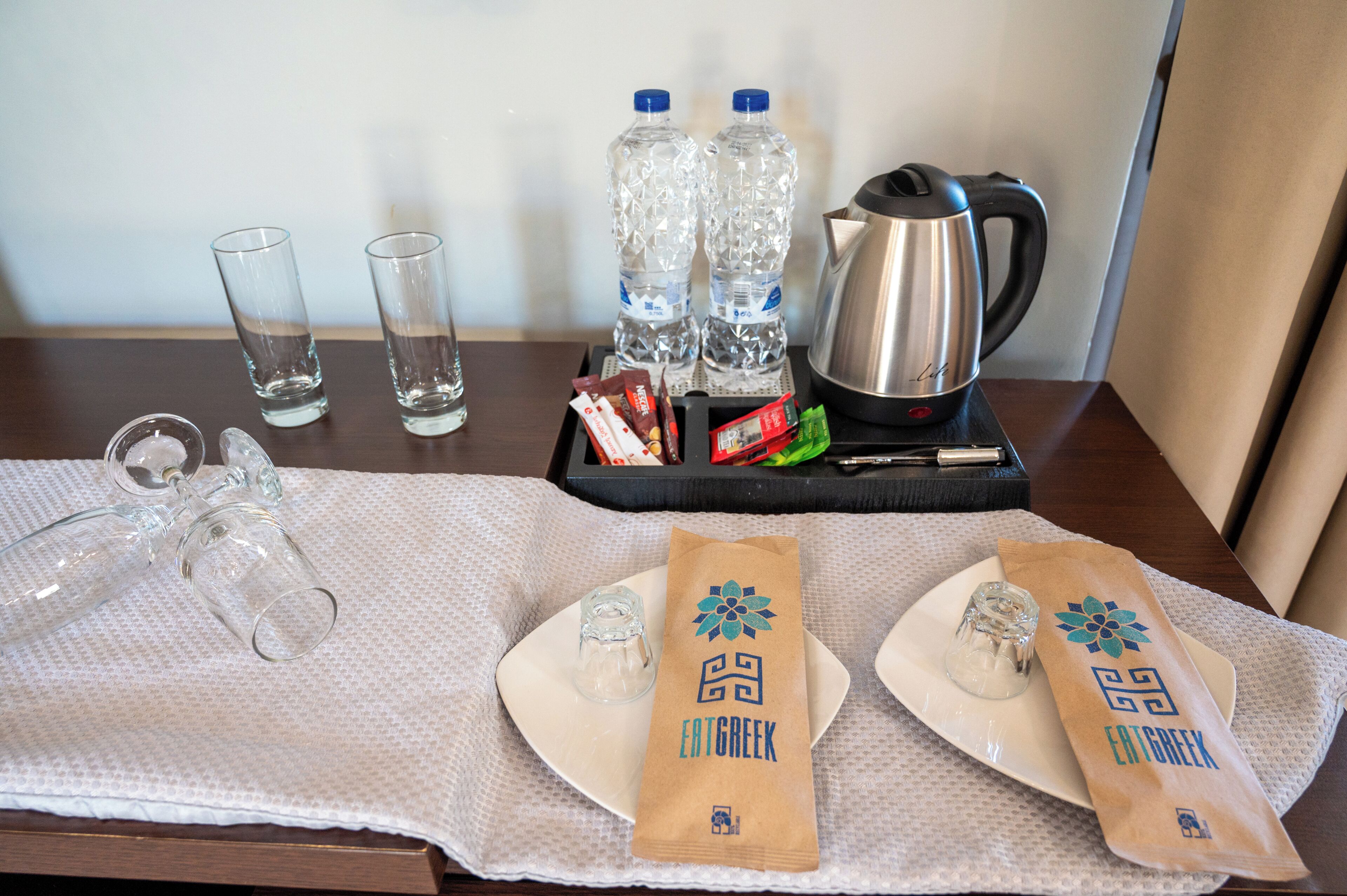 A table with a tray containing two water bottles, a kettle, and some snacks. There are also two empty glasses and a small glass on a plate.