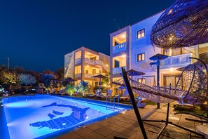 Una piscina con un diseño geométrico de azulejos, rodeada de sillones de playa y sombrillas, con un edificio que tiene balcones y una zona exterior cubierta al fondo.