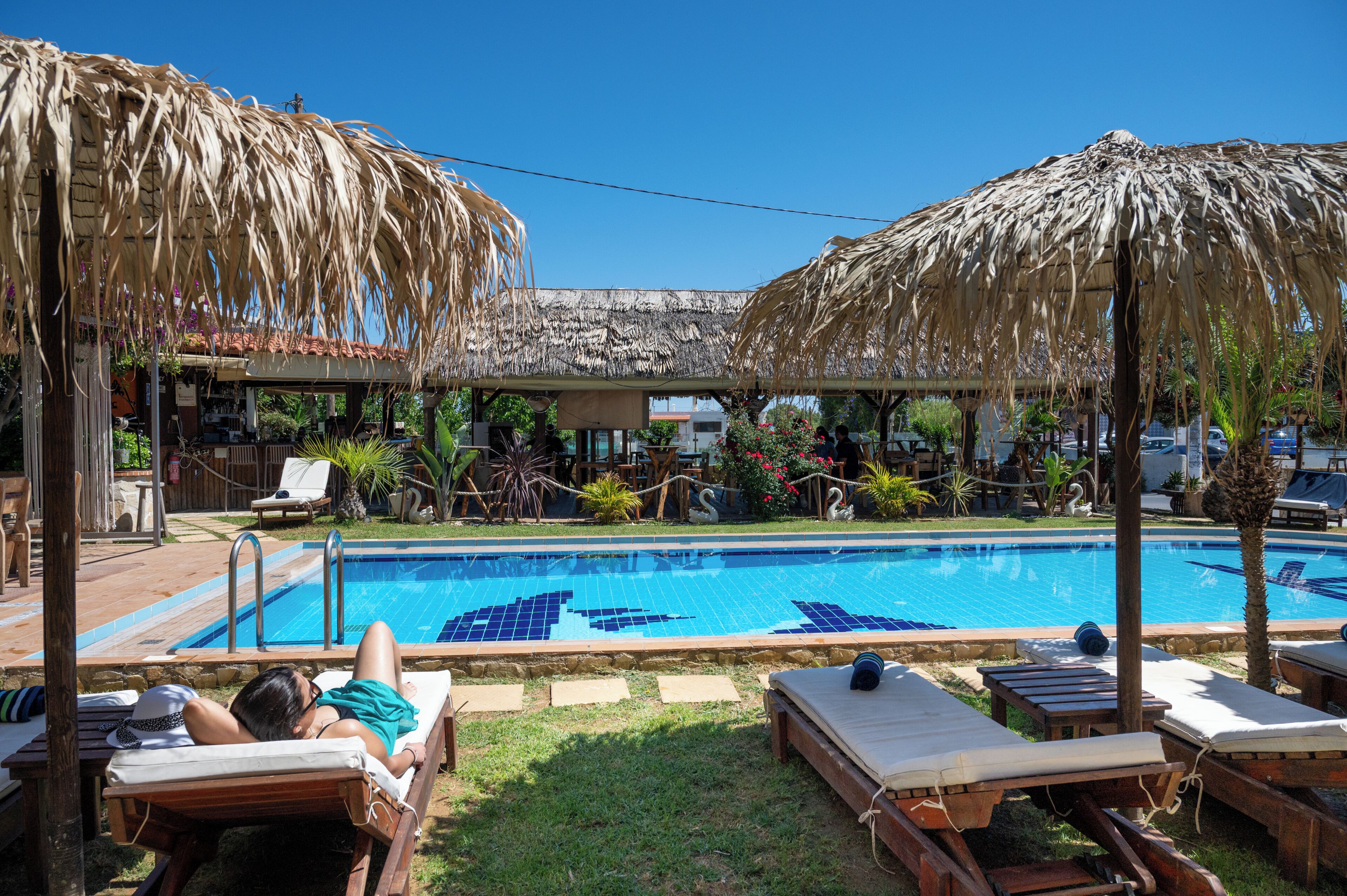 A pool area with lounge chairs and umbrellas, a clear blue sky, and a building with a tiled roof in the background.