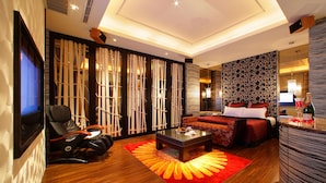 A modern hotel room with a wooden floor, a massage chair, a sofa, a bed with red bedding, and a TV.
