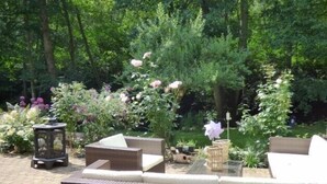 An outdoor seating area with wicker furniture and a small table, surrounded by lush greenery and flowering plants. - Wald Villa Üssbach (Alf)
