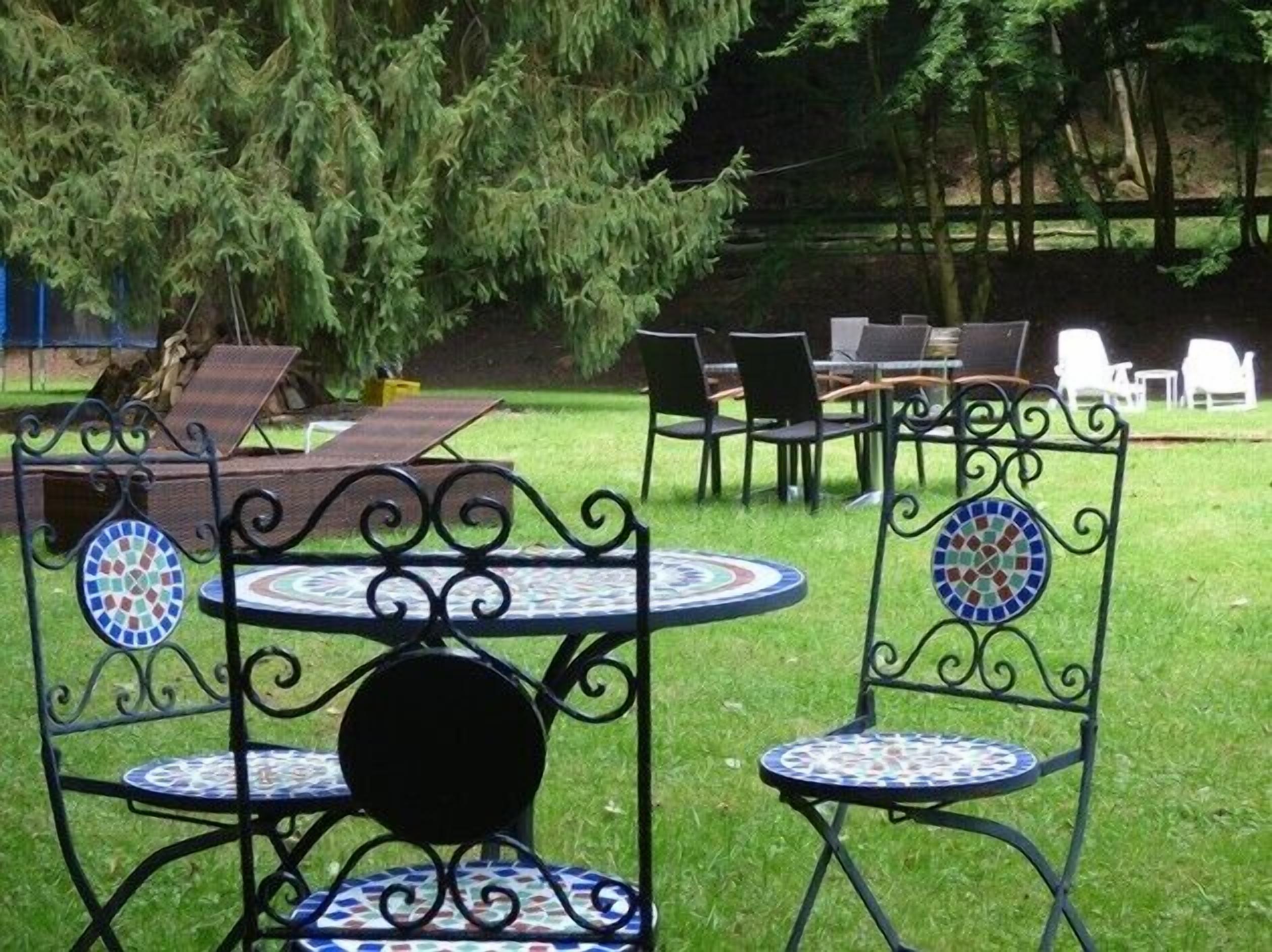Espace extérieur aménagé avec des chaises en métal et des tables rondes, entouré de pelouse et d’arbres.