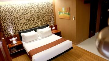 A hotel room with a neatly made bed, a patterned wallpaper, a painting on the wall, and a wooden floor.
