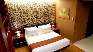 A hotel room with a large bed, wooden headboard, and a patterned wallpaper. There is a painting on the wall and a bedside table with a lamp. - Citihub Hotel @Gejayan, Yogyakarta (Yogyakarta)