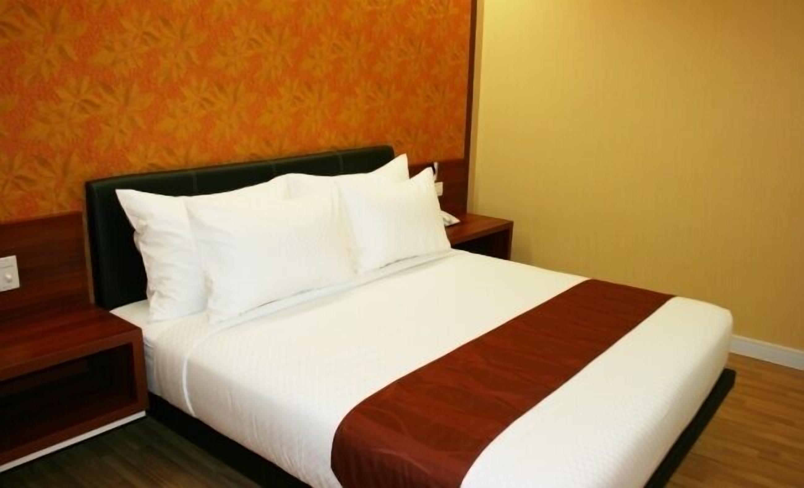 A neatly made bed with white linens and a brown accent, a wooden headboard, and a wooden nightstand with a lamp.