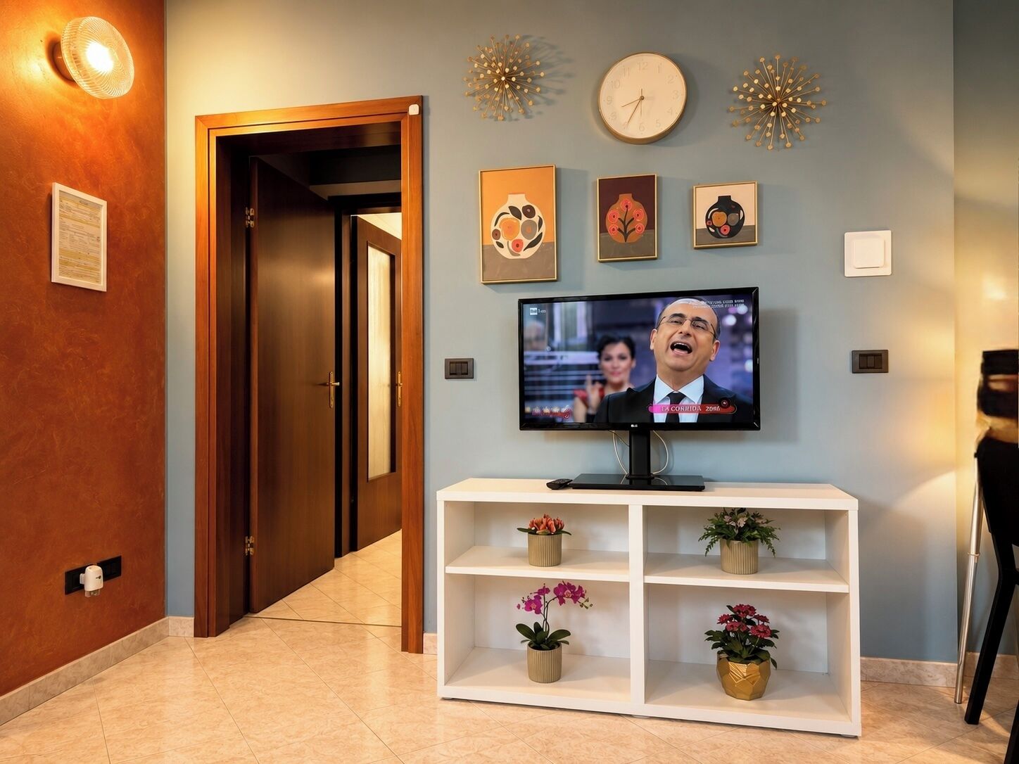 A hallway with a TV on a white console, potted plants, a clock, and decorative wall art.