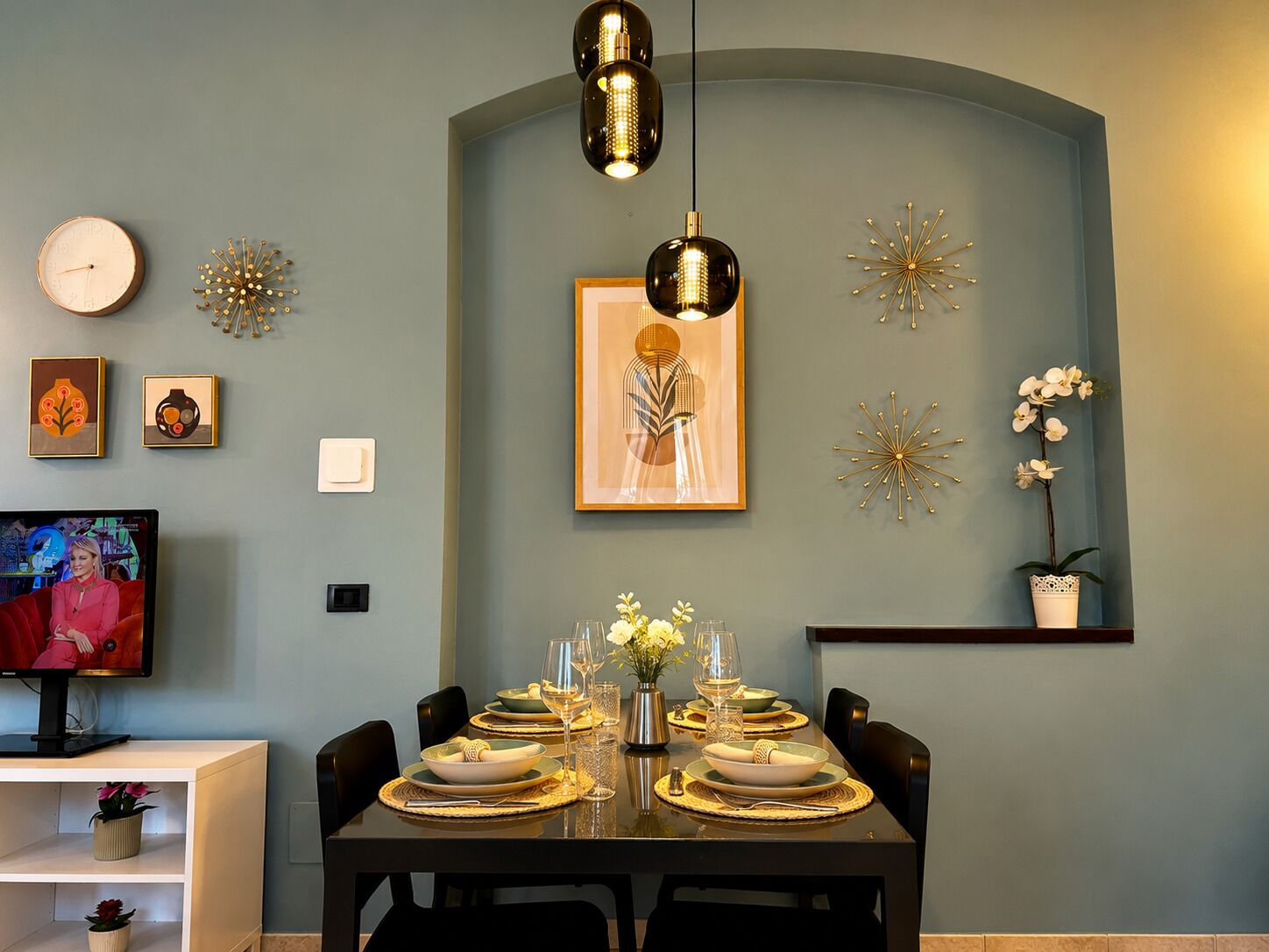 Un comedor con una mesa lista para cuatro personas, un televisor, relojes de pared y decoración mural.