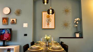 Un comedor con una mesa lista para cuatro personas, un televisor, relojes de pared y decoración mural.