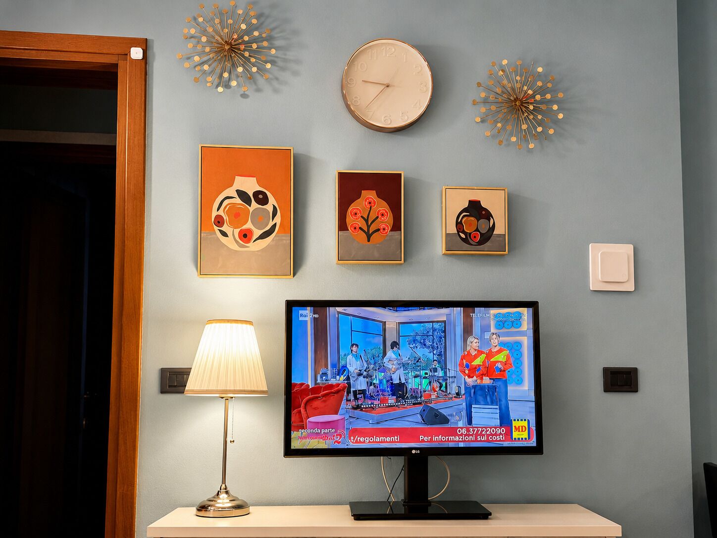 Un televisor mostrando un programa de noticias, un reloj de pared y tres obras de arte enmarcadas sobre una pared azul claro.