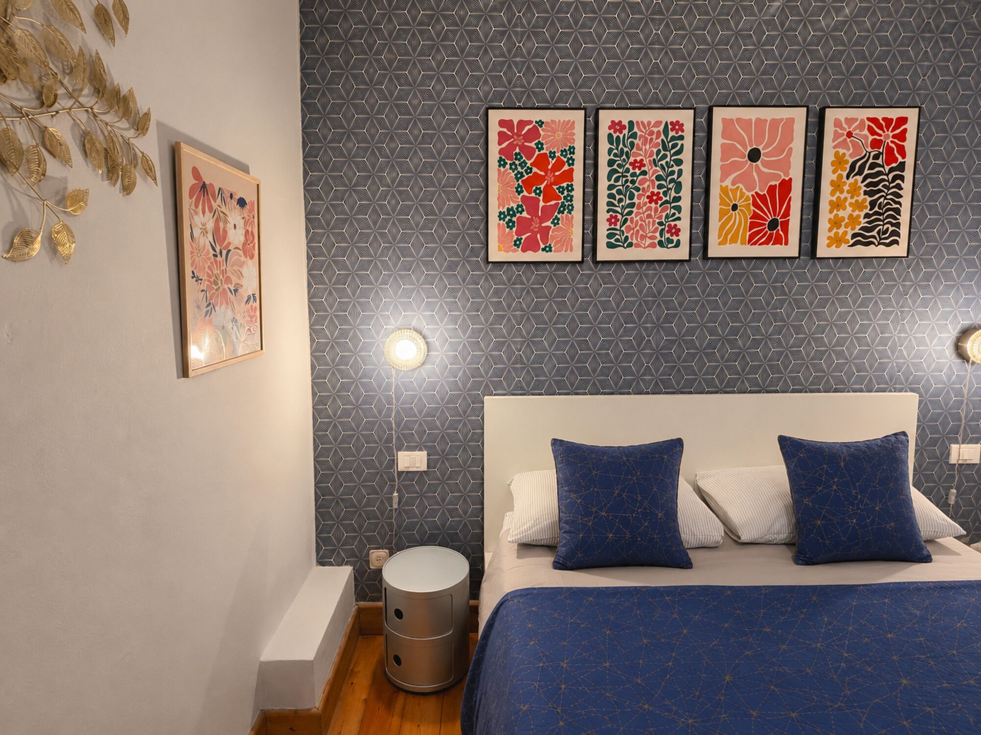 Un dormitorio con cama, almohadas, una lámpara en la pared y un cubo de basura.