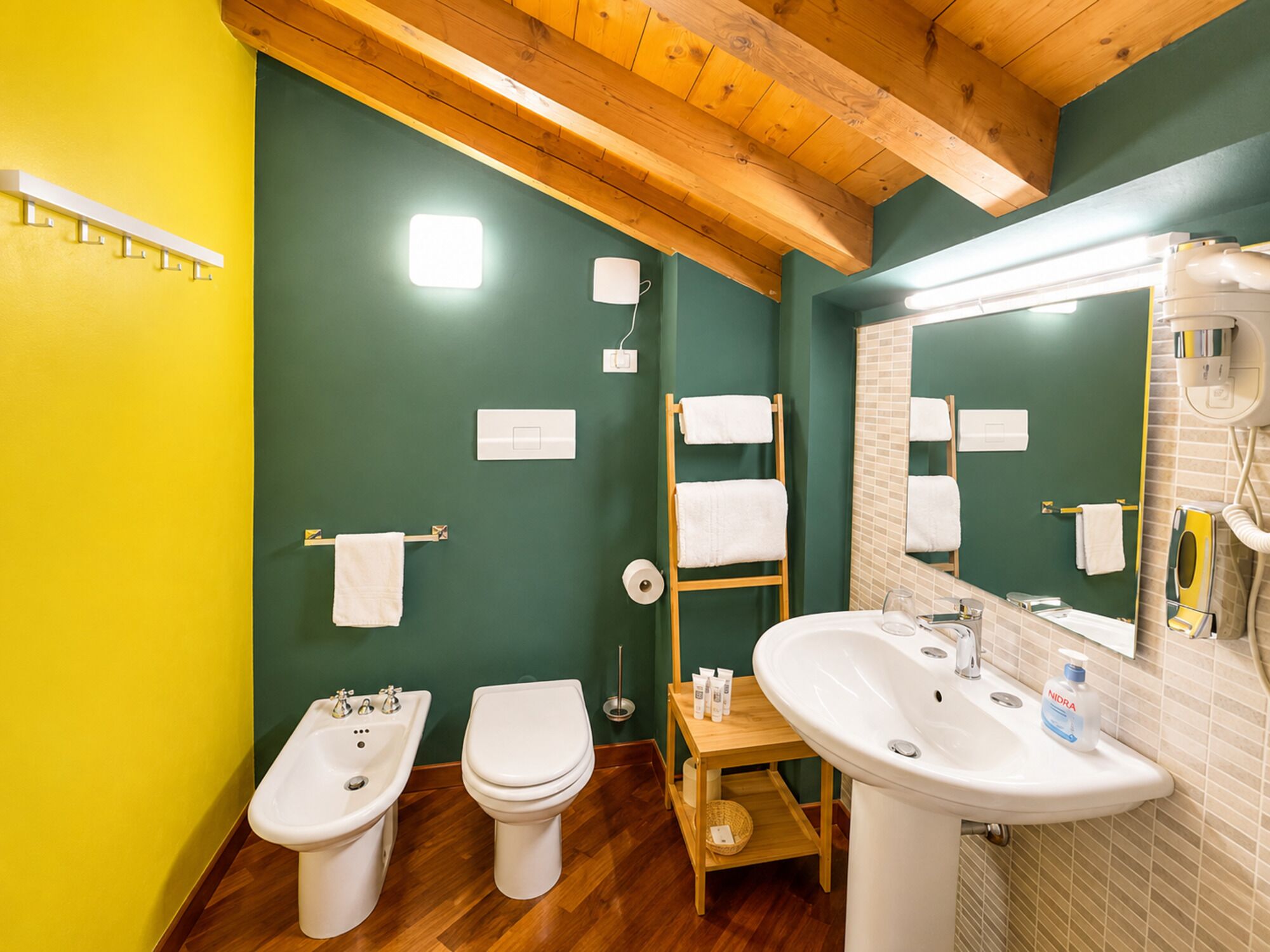 Ein Badezimmer mit Holzboden, grünen Wänden, zwei Toiletten, einem Waschbecken, einem Spiegel und einem Handtuchhalter.