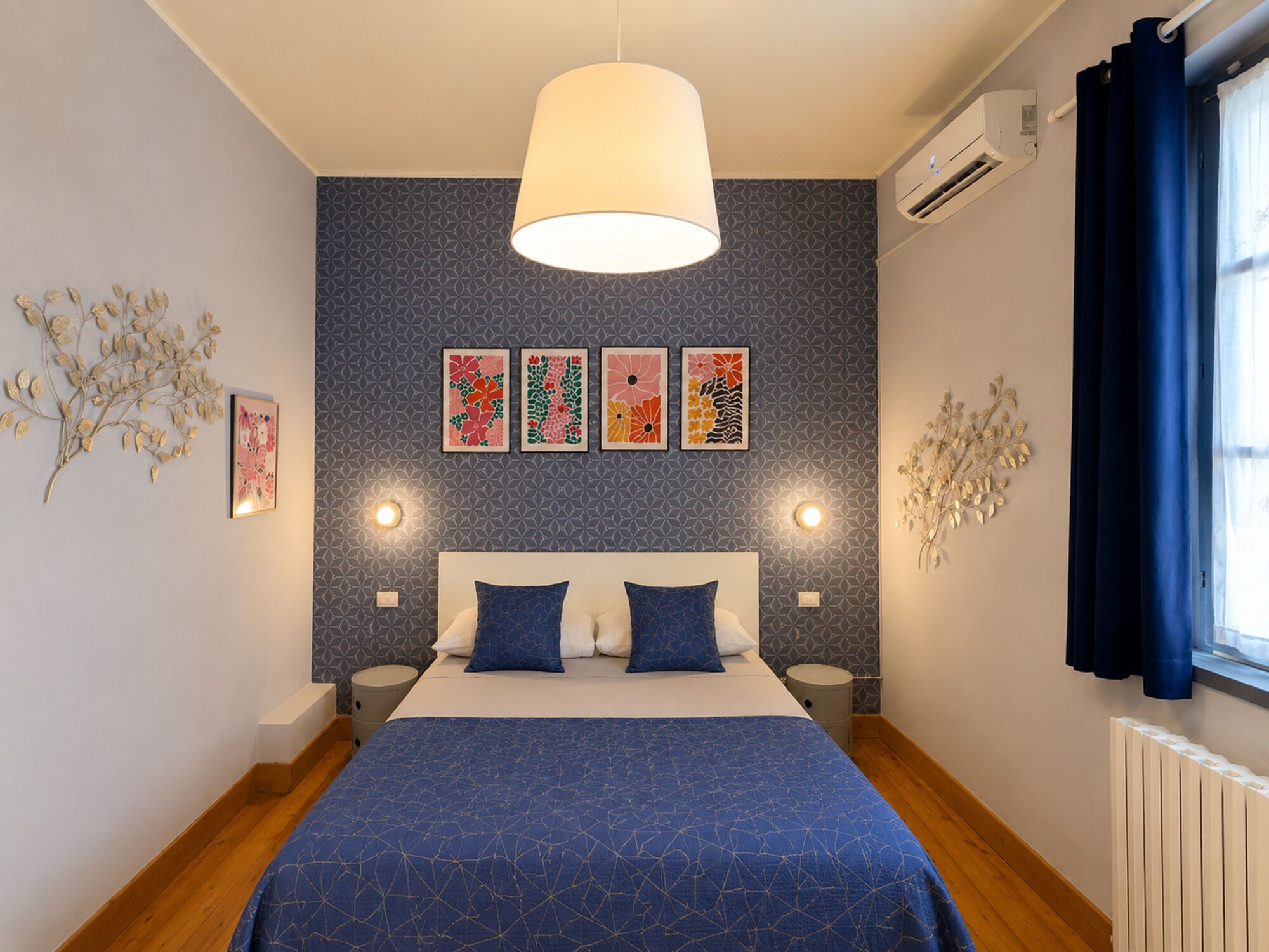 A bedroom with a bed, a hanging light fixture, wall-mounted air conditioning, and decorative wall art.