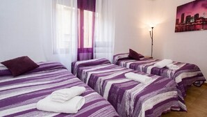 Quarto de hotel com três camas, cada uma com roupa de cama listrada em roxo e branco, uma pintura com vista para a cidade na parede e um abajur.