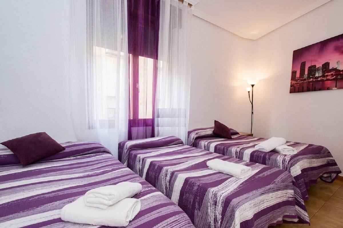 Een hotelkamer met drie bedden, elk voorzien van paars-wit gestreepte lakens, een stadsgezicht schilderij aan de muur en een vloerlamp.