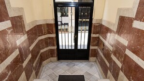 Escadaria que leva a uma porta de metal preta com portão de metal, cercada por paredes de mármore com padrões diagonais.