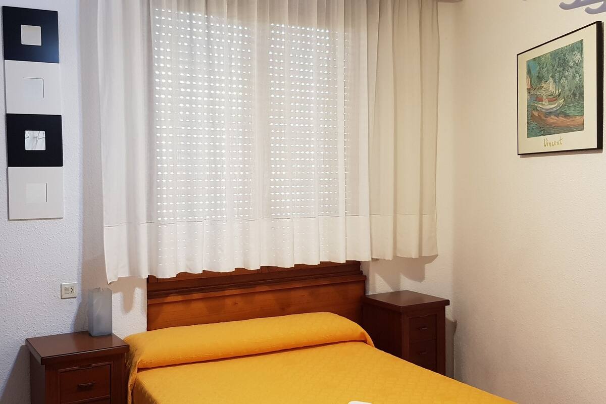 Een slaapkamer met een houten hoofdbord, een enkel bed met een oranje sprei, een nachtkastje, een lade, een wandcontactdoos en een ingelijst kunstwerk aan de muur.