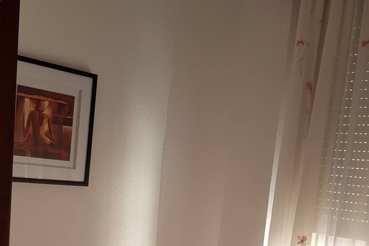 Een slaapkamer met een bed, een plank, een lijst met een afbeelding en een raam met gordijnen.