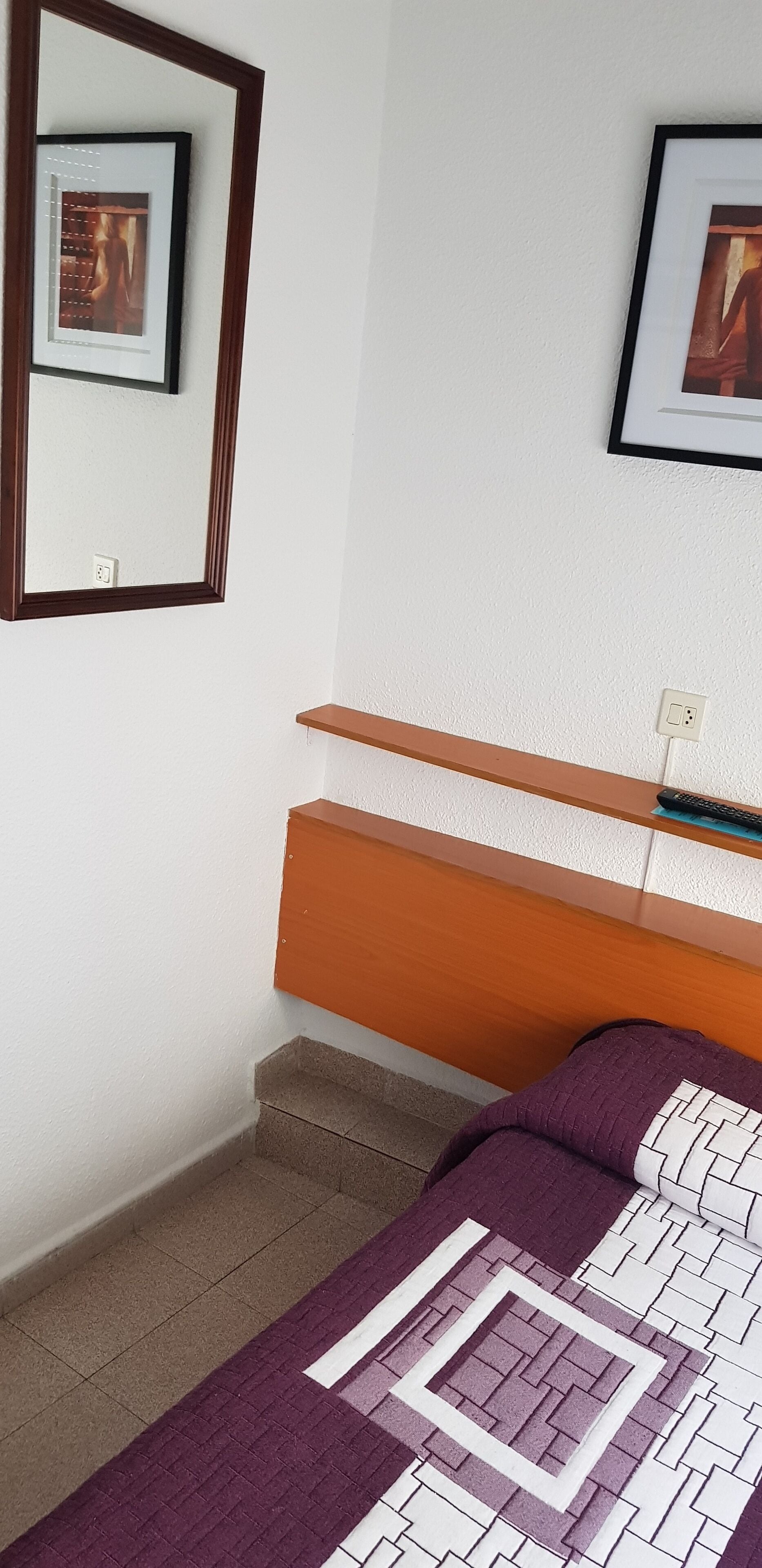 A bedroom with a bed, a wooden headboard, a framed picture on the wall, and a small shelf with a remote control.