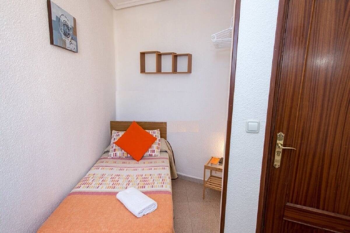 Een kleine, netjes ingerichte slaapkamer met een bed, een nachtkastje en een houten deur.