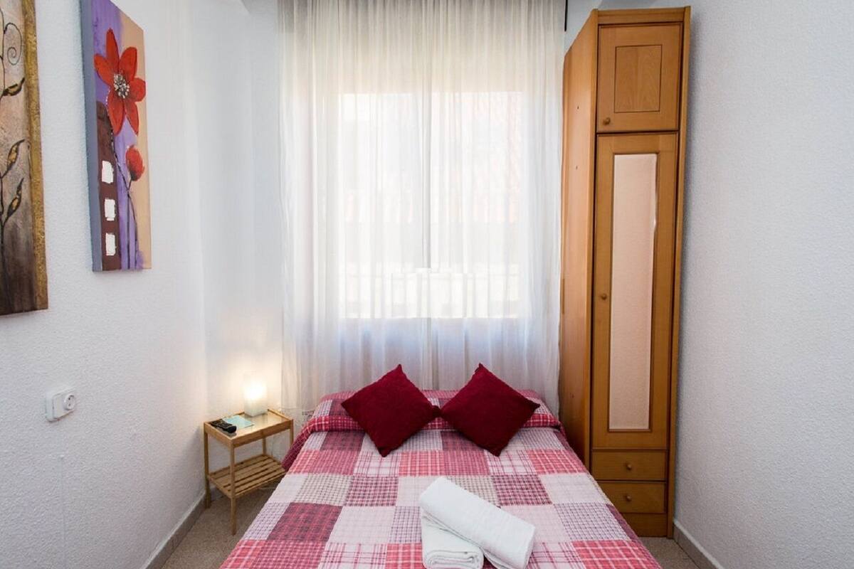 Een keurig opgemaakt bed met een geruit sprei en twee kussens, een nachtkastje met een lamp, een houten garderobe en een schilderij aan de muur.