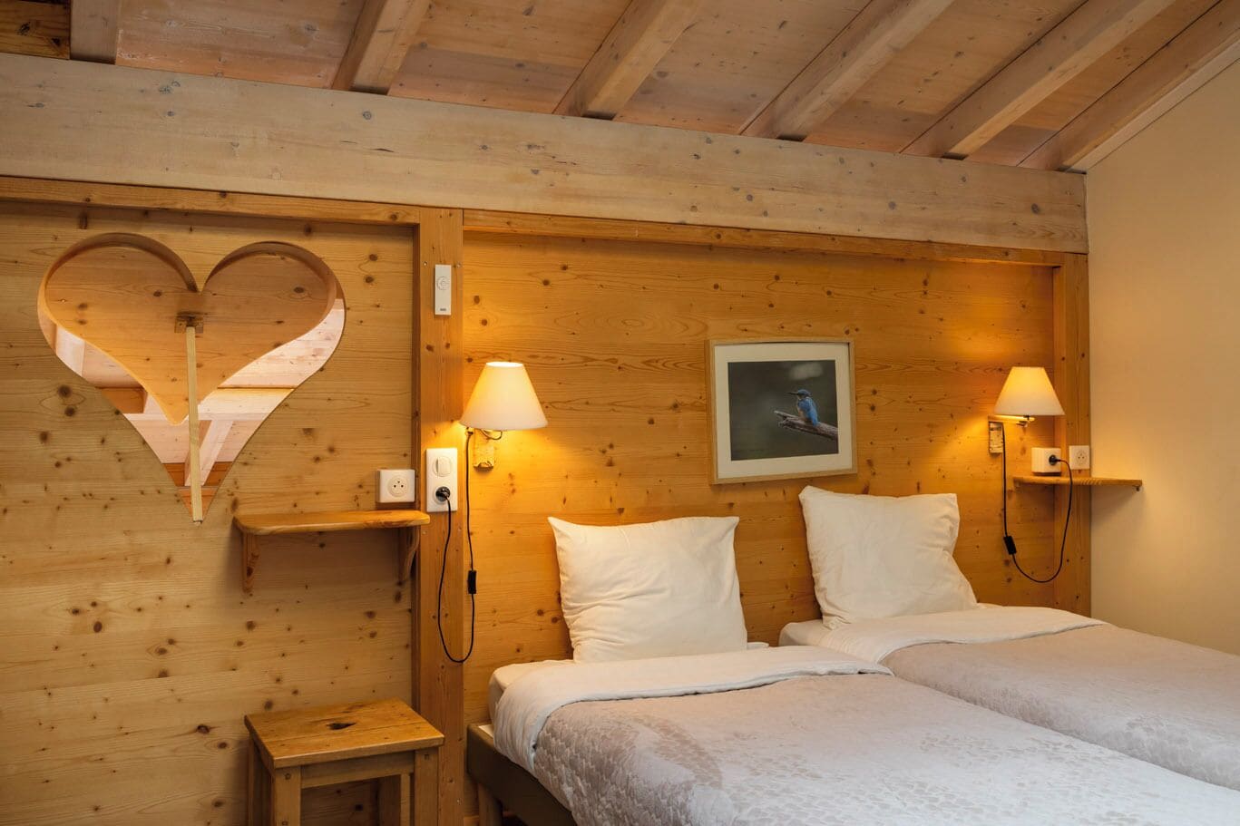 Een houten kamer met een bed, twee bedlampjes, een aan de muur hangend schilderij en een hartvormige uitsparing.