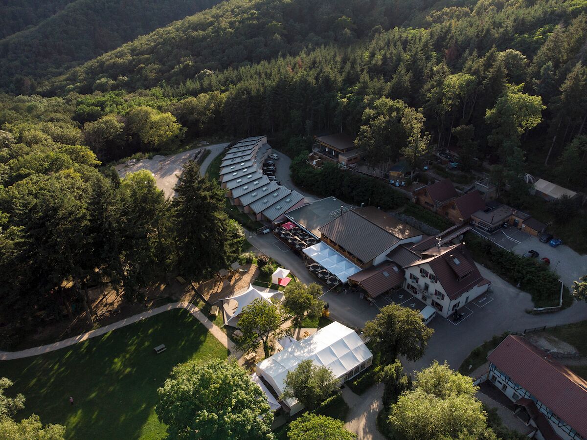 Ovenudblik over et resort med flere bygninger, en parkeringsplads og en skovklædt baggrund.