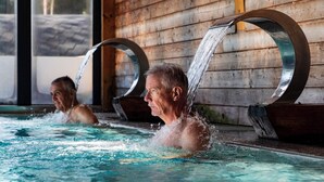 Une zone de piscine où se trouvent deux personnes dans l’eau, un mur en bois et une structure métallique d’où s’écoule de l’eau.