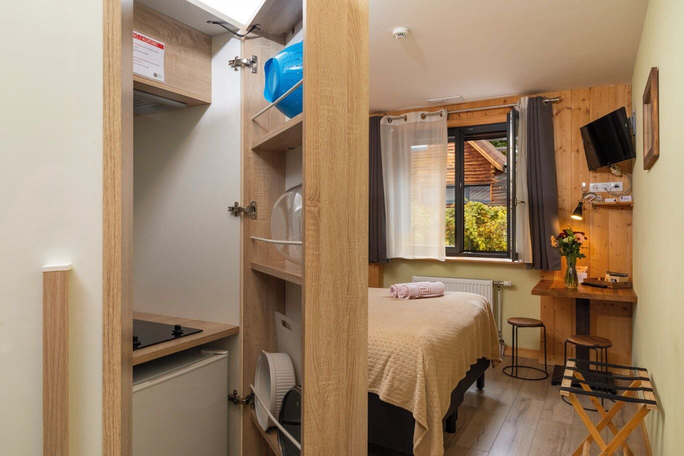 A compact bedroom with a bed, a small table, a chair, a window with curtains, and a TV mounted on the wall.