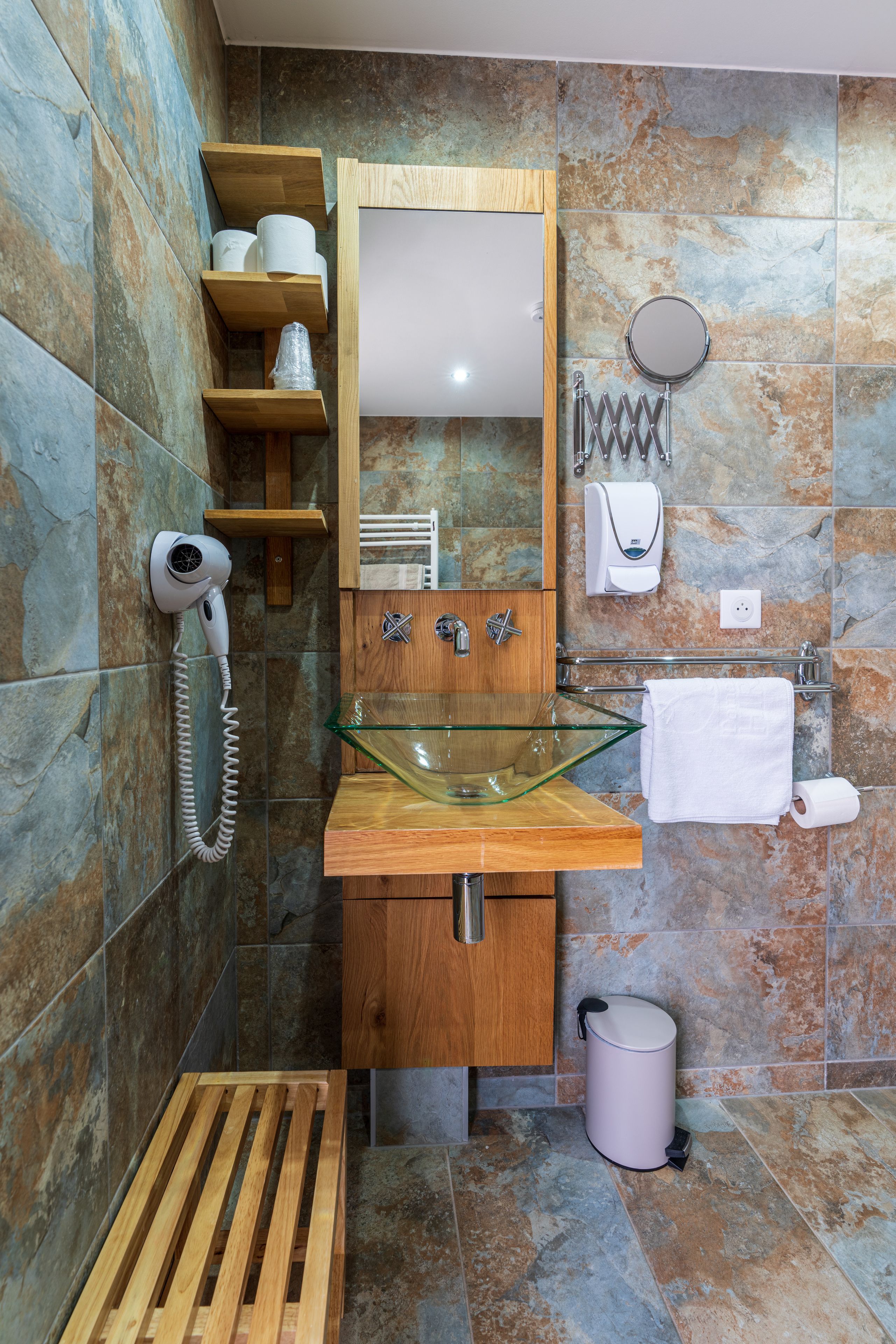 A bathroom with a glass sink, wooden shelves, a hairdryer, a towel rack, and a trash can.